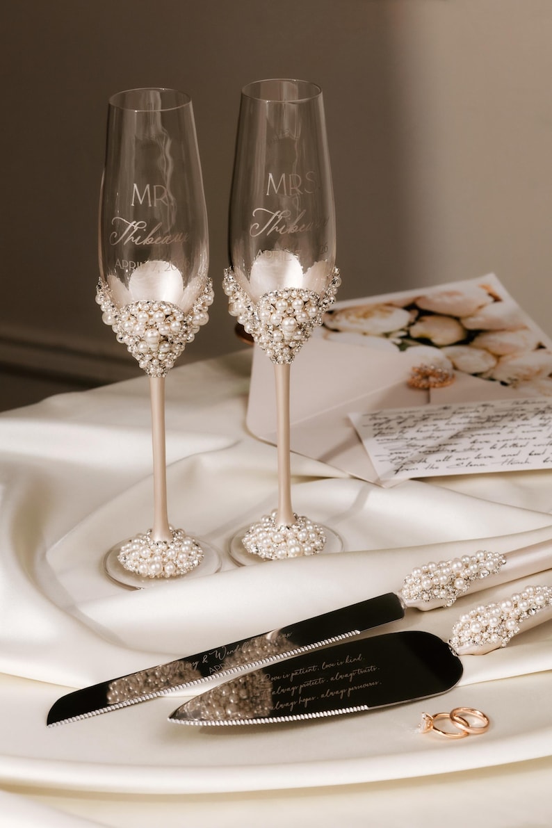 May include: Elegant champagne flutes and cake serving set with pearl embellishments. The flutes are engraved with "MR" and "MRS" and a last name. The cake server and knife have pearl accents. Wedding theme.