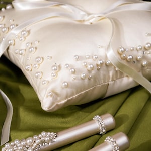 May include: Ivory satin ring bearer pillow adorned with scattered faux pearls and a satin ribbon. Two matching pearl and rhinestone embellished wands rest on a green surface. Wedding decor.