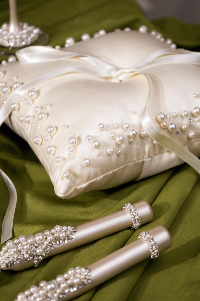 May include: Ivory satin ring bearer pillow adorned with scattered faux pearls and a satin ribbon. Two matching pearl and rhinestone embellished wands rest on a green surface. Wedding decor.