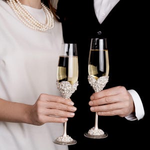 May include: Two champagne flutes filled with a golden liquid, held by a couple. The flutes have ornate pearl and rhinestone embellishments on the base and bowl. The background is black and white.
