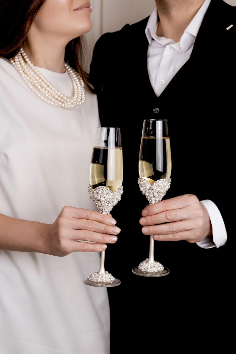 May include: Two champagne flutes filled with a golden liquid, held by a couple. The flutes have ornate pearl and rhinestone embellishments on the base and bowl. The background is black and white.