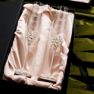 May include: Two champagne flutes with pearl and rhinestone embellishments, presented in a black gift box. The flutes have the inscription "Mrs. Entygen". The flutes are displayed on a satin fabric.