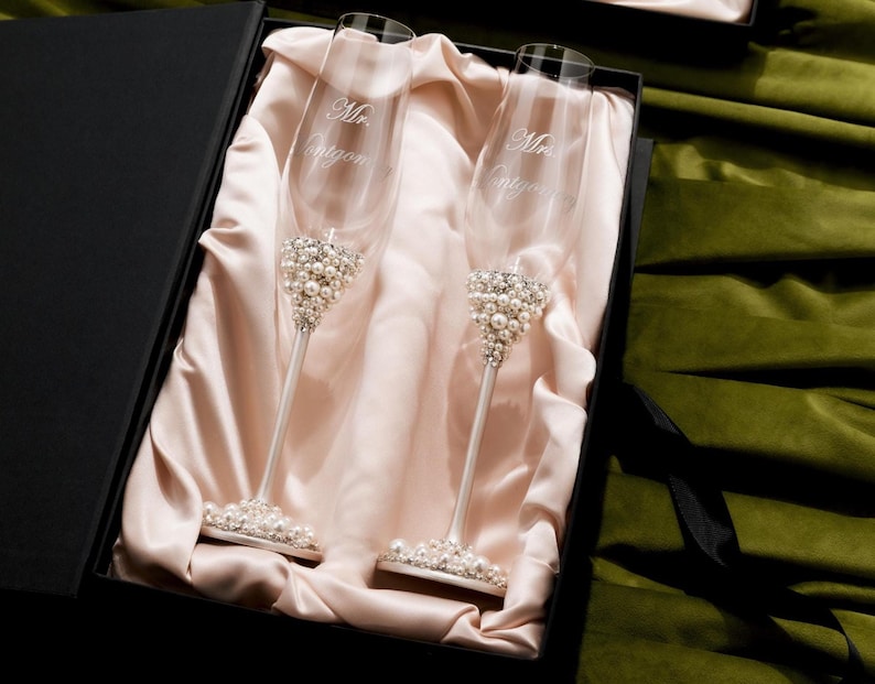 May include: Two champagne flutes with pearl and rhinestone embellishments, presented in a black gift box. The flutes have the inscription "Mrs. Entygen". The flutes are displayed on a satin fabric.