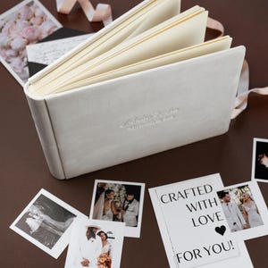 May include: A light gray, open photo album with cream-colored pages and a ribbon. Several printed photos and a card with the text "CRAFTED WITH LOVE FOR YOU!" are scattered around the album.