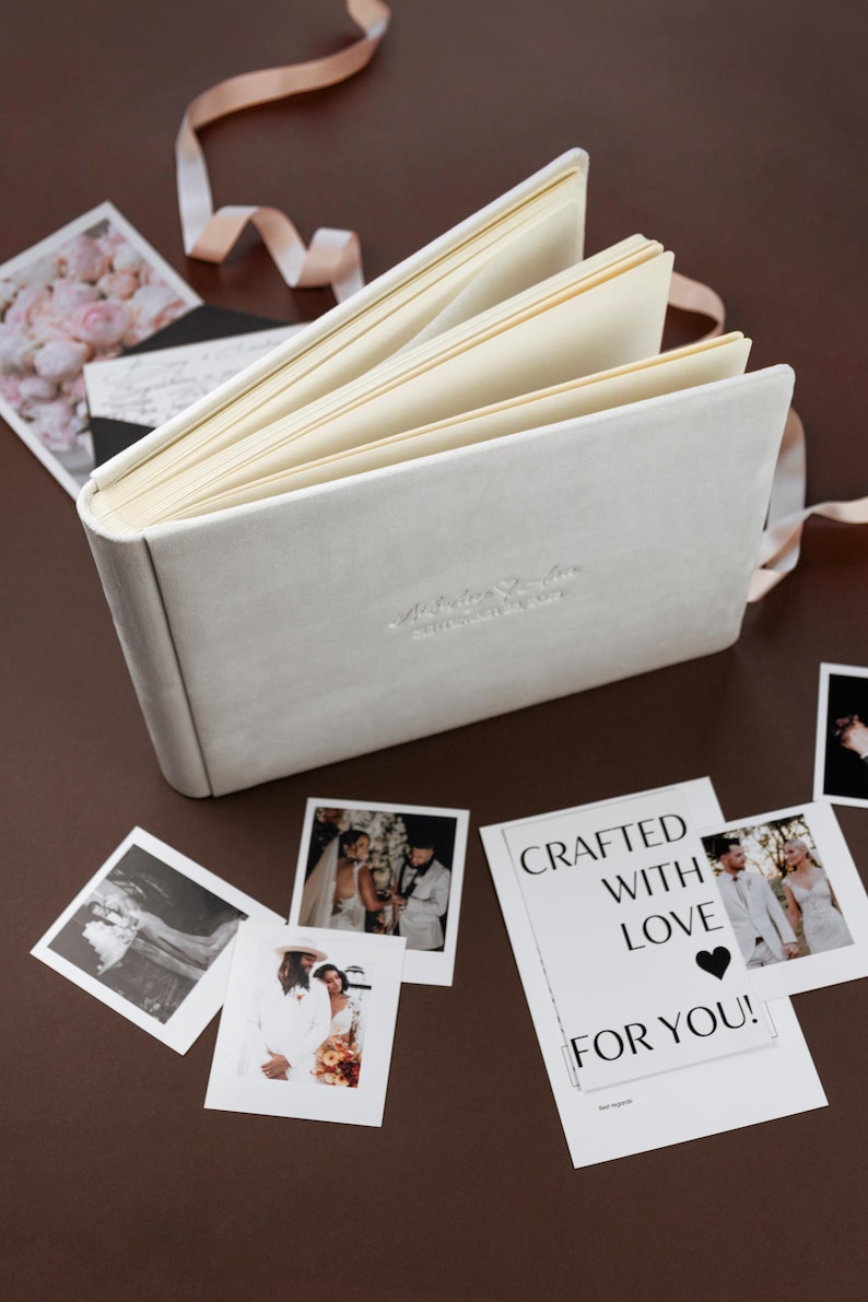 May include: A light gray, open photo album with cream-colored pages and a ribbon. Several printed photos and a card with the text "CRAFTED WITH LOVE FOR YOU!" are scattered around the album.