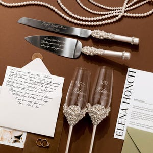 May include: A wedding cake knife and server set with pearl accents, along with two champagne flutes. The flutes are engraved with "MRS" and "MR" and the couple's last name and wedding date. A handwritten note and wedding rings are also present.