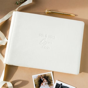 May include: A white wedding photo album with embossed text reading "MR & MRS Byren 3.10.23" sits on a neutral surface. Two printed photos and a gold pen with a diamond-shaped top are also present. A white ribbon and greenery add to the composition.