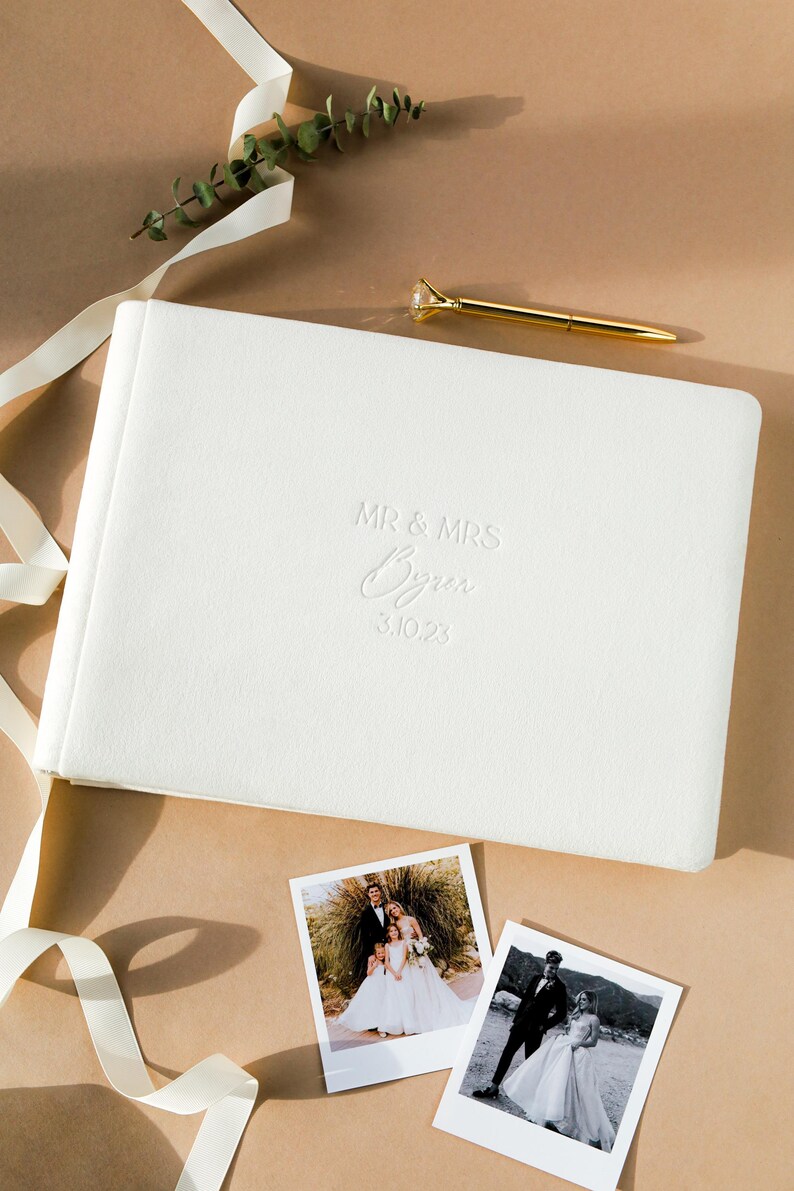 May include: A white wedding photo album with embossed text reading "MR & MRS Byren 3.10.23" sits on a neutral surface. Two printed photos and a gold pen with a diamond-shaped top are also present. A white ribbon and greenery add to the composition.