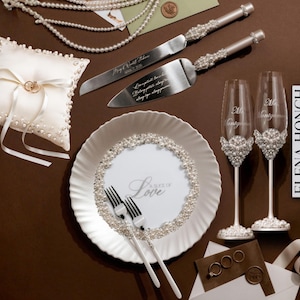 May include: Elegant wedding accessories set on a brown surface. Includes a ring pillow, cake server and knife, champagne flutes, a plate with "Love" text, and pearl accents. The set features a white and silver color scheme.