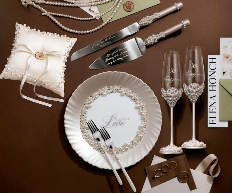 May include: Elegant wedding accessories set on a brown surface. Includes a ring pillow, cake server and knife, champagne flutes, a plate with "Love" text, and pearl accents. The set features a white and silver color scheme.