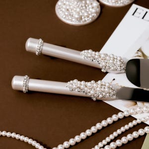May include: Elegant wedding cake server and knife set with pearl and rhinestone embellishments. The handles are white, and the blades are silver. The set is displayed with pearl strands and two champagne flutes with pearl bases.