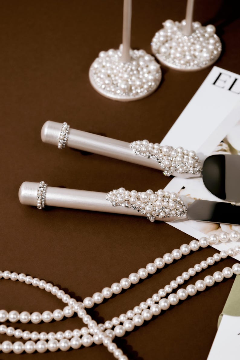 May include: Elegant wedding cake server and knife set with pearl and rhinestone embellishments. The handles are white, and the blades are silver. The set is displayed with pearl strands and two champagne flutes with pearl bases.
