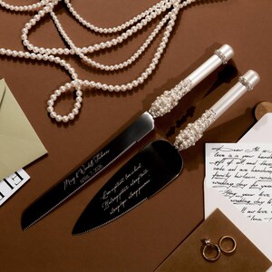 May include: A wedding cake knife and server set with pearl and rhinestone accents. The handles are white, and the blades are black. The knife and server have engraved text. A strand of pearls and wedding rings are also in the image.