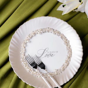 May include: A white plate with a pearl and rhinestone border, two forks, and the text "A Slice of Love". The plate is on a green cloth, with a white lily flower in the background.