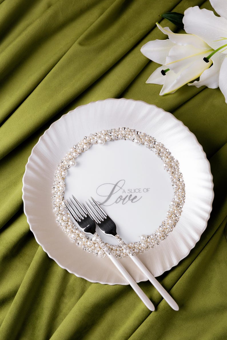 May include: A white plate with a pearl and rhinestone border, two forks, and the text "A Slice of Love". The plate is on a green cloth, with a white lily flower in the background.