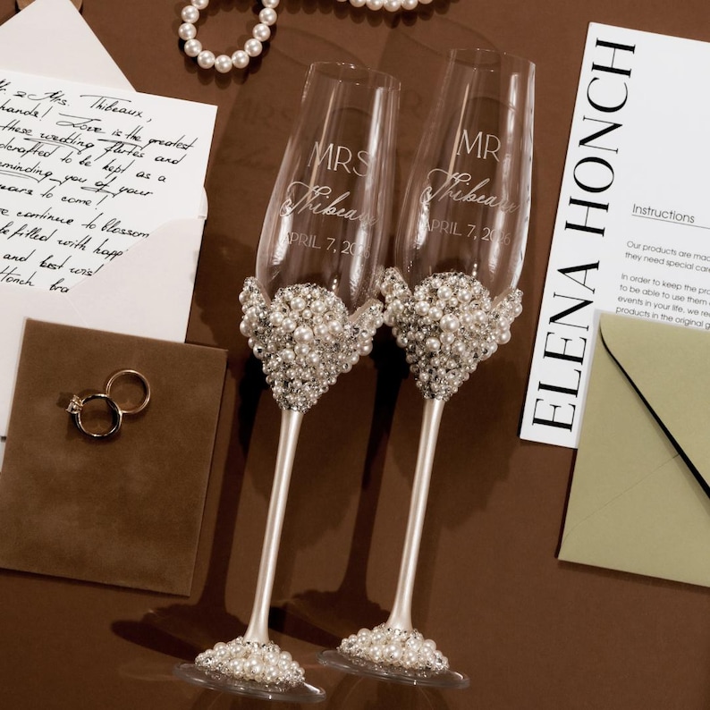 May include: Two champagne flutes with pearl embellishments. The flutes are engraved with "MRS" and "MR", names, and the date "APRIL 7, 2016". The glasses sit on a brown surface with wedding-related items.