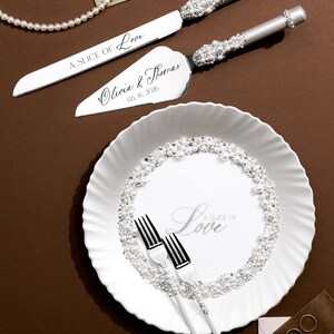 May include: A wedding cake knife and server set with silver handles and crystal accents. The knife and server are engraved with "A SLICE OF Love" and the names "Olivia & Thomas" with the date "05.16.2026". A white plate with "A SLICE OF Love" text and pearl accents.