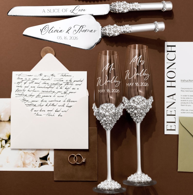 May include: A wedding-themed still life featuring a cake knife and server set, two champagne flutes, and a handwritten letter. The knife and server are silver with crystal accents. The flutes are adorned with crystals and personalized with names and a date.