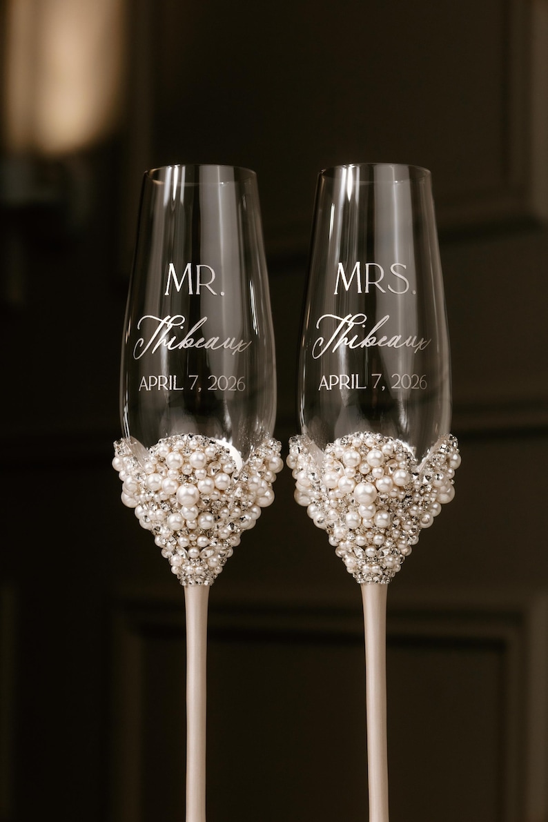 May include: Two champagne flutes with pearl and crystal embellishments at the base. The flutes are engraved with "MR. Thibeau" and "MRS. Thibeau" and the date "APRIL 7, 2026". The stems are a light cream color.