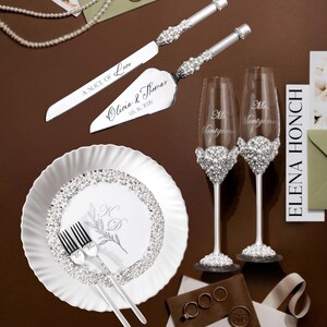 May include: Elegant wedding set featuring a cake knife and server, two champagne flutes, a plate, and forks. The set is adorned with sparkling embellishments. The cake server is engraved with "A Slice of Love" and the names "Olivia & Thomas" and the date "05.16.2026".
