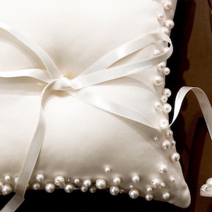 May include: Ivory satin ring pillow adorned with scattered pearls around the edges. A white satin ribbon is tied in a bow on the front. Wedding accessory.