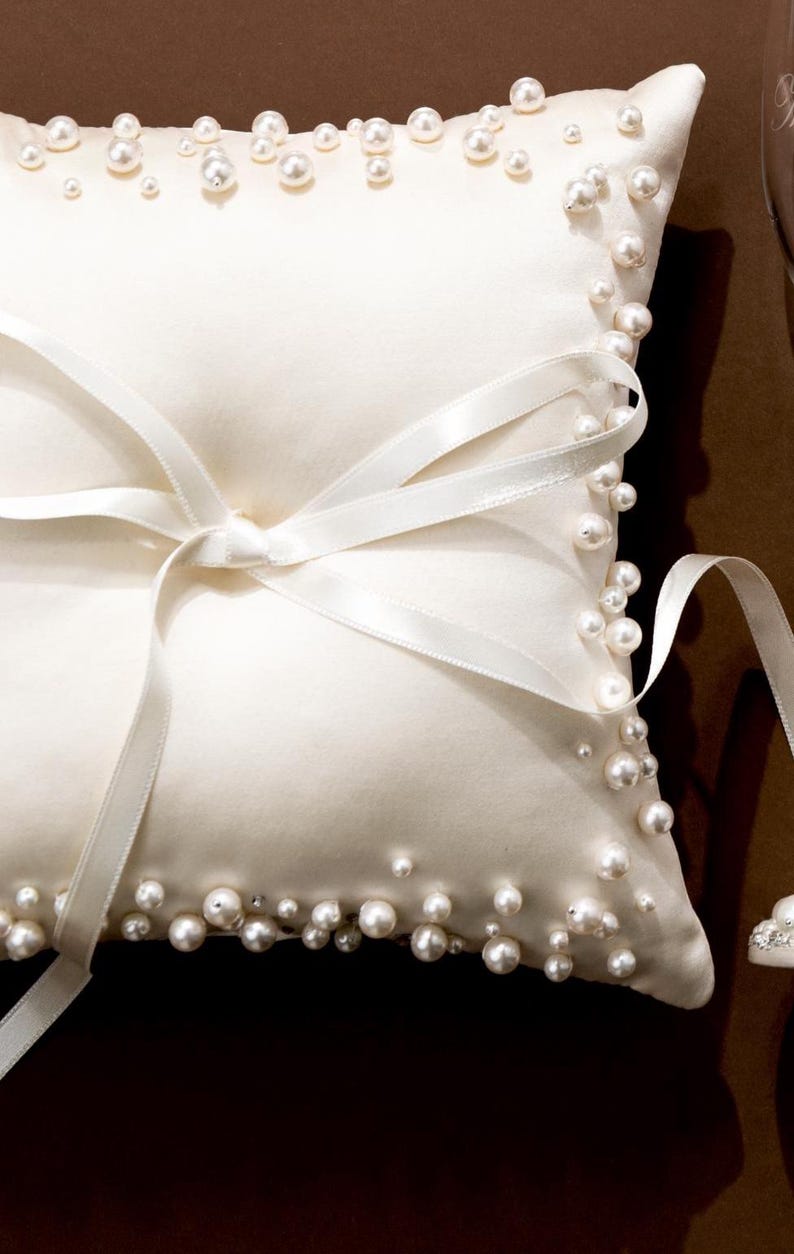 May include: Ivory satin ring pillow adorned with scattered pearls around the edges. A white satin ribbon is tied in a bow on the front. Wedding accessory.