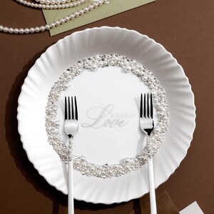 May include: A white plate with a pearl and crystal border, two forks, and the word "Love" in script. The plate is surrounded by pearls, a photo, and wedding rings, suggesting a wedding theme.