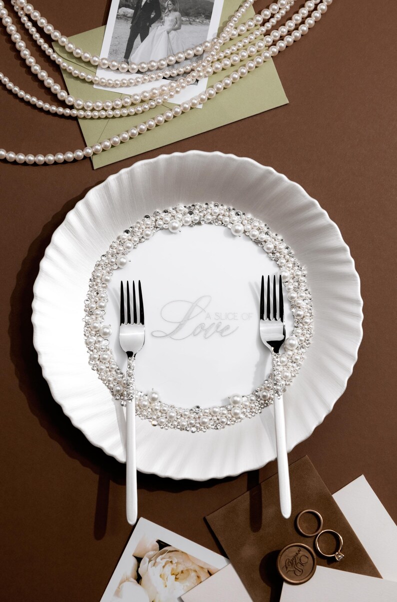 May include: A white plate with a pearl and crystal border, two forks, and the word "Love" in script. The plate is surrounded by pearls, a photo, and wedding rings, suggesting a wedding theme.