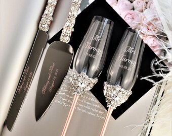 May include: Rose gold wedding cake knife and server set with crystal embellishments and a matching set of champagne flutes with crystal embellishments. The knife and server are engraved with the names "Hillary and Klint" and the date "August 13, 2022". The champagne flutes are engraved with "Mr. Turney 08.13.22" and "Mrs. Turney 08.13.22".