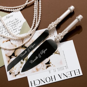 May include: A wedding cake knife and server set with black blades and pearl-adorned handles. The set is displayed with a pearl necklace and a card that reads "Camille & Ryan".