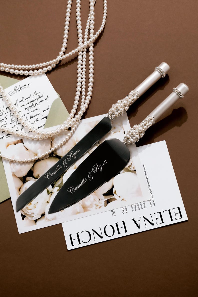 May include: A wedding cake knife and server set with black blades and pearl-adorned handles. The set is displayed with a pearl necklace and a card that reads "Camille & Ryan".