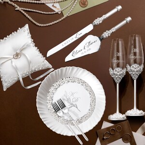 May include: A collection of wedding accessories, including a white ring pillow, cake server and knife set, champagne flutes, and a decorative plate. The cake server and knife are engraved with the words "A Slice of Love" and the couple's names and date.