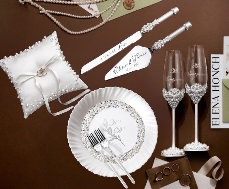 May include: A collection of wedding accessories, including a white ring pillow, cake server and knife set, champagne flutes, and a decorative plate. The cake server and knife are engraved with the words "A Slice of Love" and the couple's names and date.