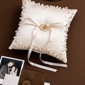 May include: Ivory satin ring pillow adorned with faux pearls and a white ribbon. Two gold wedding rings rest on the pillow. A strand of pearls and wedding stationery are also visible on a brown surface.