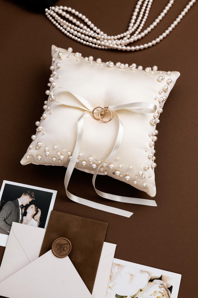 May include: Ivory satin ring pillow adorned with faux pearls and a white ribbon. Two gold wedding rings rest on the pillow. A strand of pearls and wedding stationery are also visible on a brown surface.