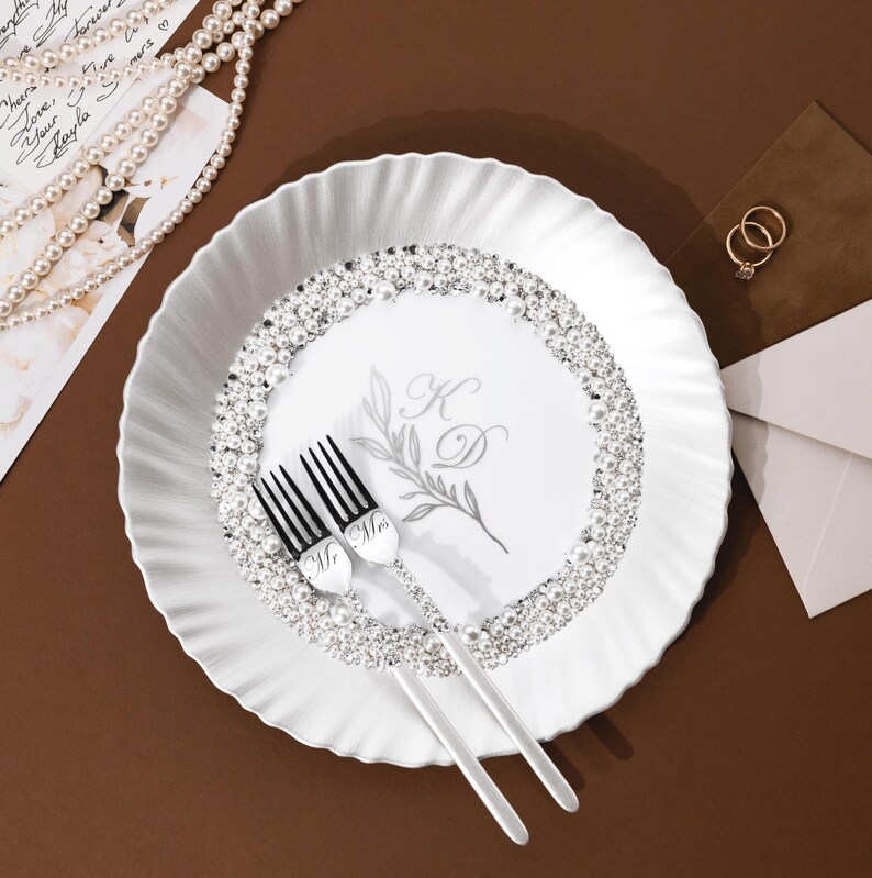 May include: A white decorative plate with a ruffled edge, adorned with a ring of faux pearls and rhinestones. Two forks with "Mr" and "Mrs" engravings rest on the plate. The plate features a floral design and initials.