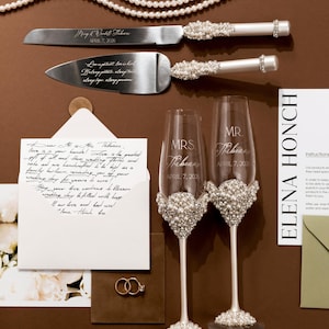 May include: Wedding cake knife and server set with pearl and rhinestone accents. Includes two champagne flutes with pearl embellishments, and the names "Mrs. Thibeau" and "Mr. Thibeau" engraved on them. Also includes wedding rings and a handwritten note.
