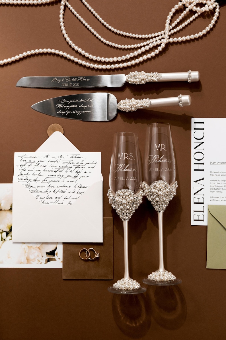May include: Wedding cake knife and server set with pearl and rhinestone accents. Includes two champagne flutes with pearl embellishments, and the names "Mrs. Thibeau" and "Mr. Thibeau" engraved on them. Also includes wedding rings and a handwritten note.