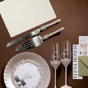 May include: Wedding-themed set featuring a cream-colored guest book, cake knife and server, two champagne flutes, and a decorative plate. The knife and server have pearl accents. The plate and flutes are decorated with pearls. The plate has the text "A Slice of Love".