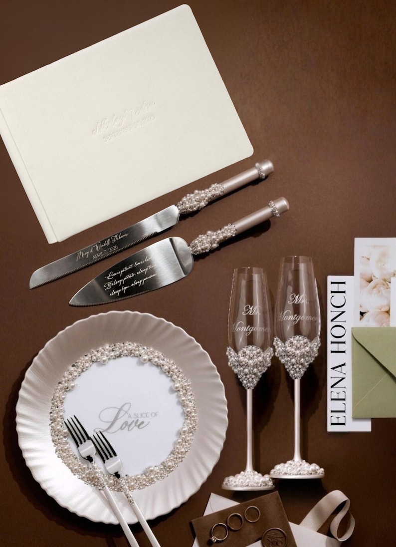 May include: Wedding-themed set featuring a cream-colored guest book, cake knife and server, two champagne flutes, and a decorative plate. The knife and server have pearl accents. The plate and flutes are decorated with pearls. The plate has the text "A Slice of Love".