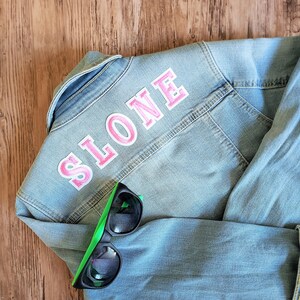 May include: A light blue denim jacket with a pink "SLONE" patch on the back. A pair of black sunglasses with green frames are lying on top of the jacket.