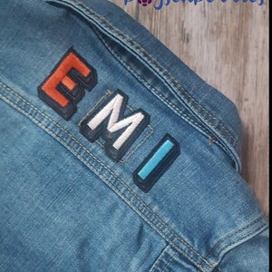 Puede incluir: Una chaqueta vaquera azul con la palabra "EMI" escrita en parches de tela de colores.