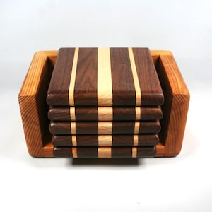 May include: A set of six square wooden coasters with alternating dark brown and light wood stripes, stacked in a light brown wooden holder. The coasters are approximately 4 inches square.