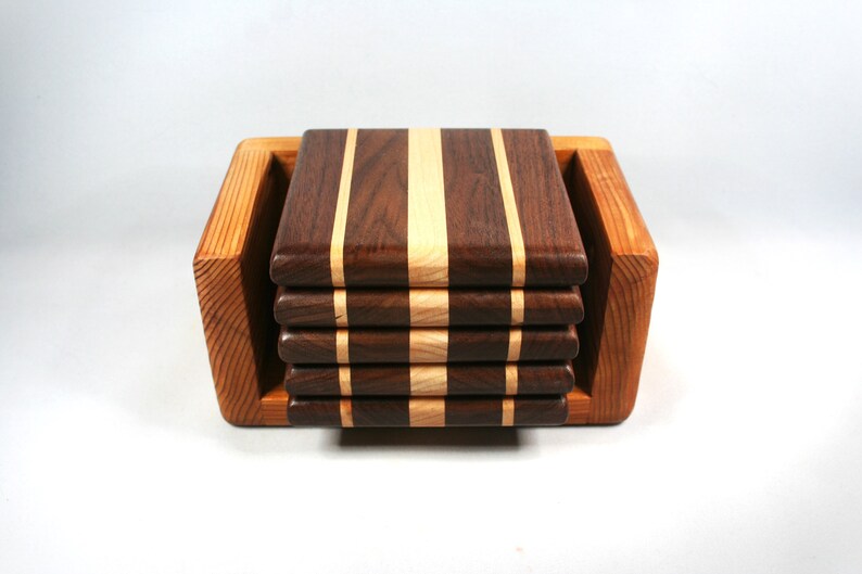 May include: A set of six square wooden coasters with alternating dark brown and light wood stripes, stacked in a light brown wooden holder. The coasters are approximately 4 inches square.