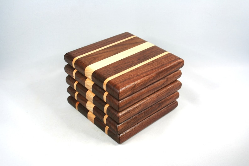 May include: A stack of five square wooden coasters. Each coaster is made of dark brown wood with a light-colored wood stripe running across the center. The coasters are stacked neatly on a white background.