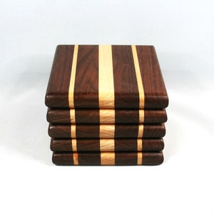May include: A stack of five square wooden coasters. Each coaster is made of dark brown wood with a light-colored wood stripe down the center and on each side. The coasters are stacked neatly.
