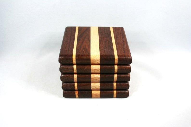 May include: A stack of five square wooden coasters. Each coaster is made of dark brown wood with a light-colored wood stripe down the center and on each side. The coasters are stacked neatly.