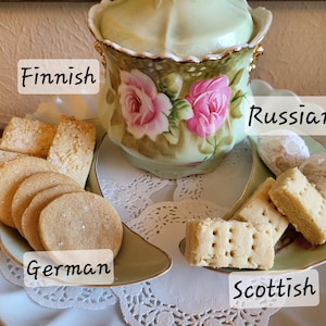 May include: A light green and gold floral tea set with a lid, surrounded by various cookies on a decorative plate. The cookies are labeled as Finnish, German, Russian, and Scottish.
