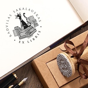 Puede incluir: Un sello de madera con un diseño decorativo y una cinta marrón, presentado en una caja de cartón. La impresión del sello presenta un gato negro sobre libros, con el texto "RADVILAS SAKALAUSKAS EX LIBRIS". Una pluma estilográfica descansa cerca.