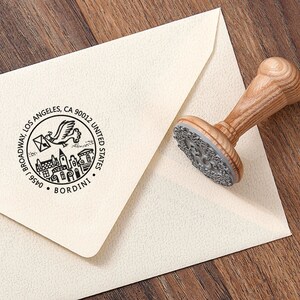 May include: A wooden stamp with a detailed design, including a flying bird carrying an envelope, buildings, and the text "0456 J BROADWAY, LOS ANGELES, CA 90012 UNITED STATES. BORDINI" is next to a cream-colored envelope.